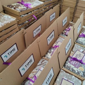 A large volume of Branded Corporate BakeBox Gifts with Time Investments Logo on the inside label with a message saying "Time to enjoy a treat on us". The BakeBox contains Chocolate Brownies, Caramel Shortcake and Scottish Tablet in cellophane tied with a purple ribbon.